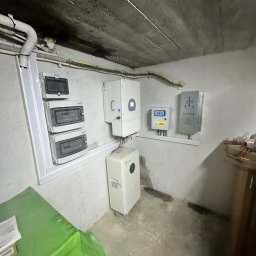 Profitenergy Spółka z ograniczoną odpowiedzialnością - Instalacja urządzeń w pomieszczeniu piwnicznym: falownik, licznik energii, rozdzielnica, akumulator, okablowanie. Widoczny surowy betonowy sufit i ślady wilgoci na ścianach.