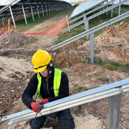 Profitenergy Spółka z ograniczoną odpowiedzialnością - Pracownik w kasku i kamizelce odblaskowej, w czerwonych rękawicach, przywiązuje metalową konstrukcję pod panele słoneczne na tle częściowo zamontowanej instalacji na gruncie.