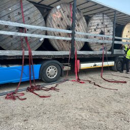 Profitenergy Spółka z ograniczoną odpowiedzialnością - Transport kabli na drewnianych bębnach, zabezpieczonych czerwonymi pasami, na platformie ciężarówki z niebieską kabiną, widok z boku, na żwirowym podłożu.