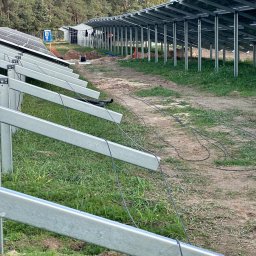 Profitenergy Spółka z ograniczoną odpowiedzialnością - Montaż konstrukcji wsporczej dla paneli słonecznych na terenie zielonym, widoczne kable i tymczasowa toaleta TOI TOI w tle.