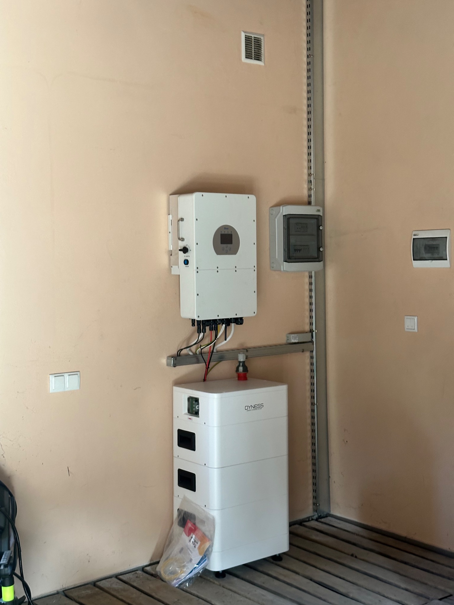 Biały inwerter i moduł bateryjny Dyness zamontowane na ścianie w pomieszczeniu. Widoczne przewody, puszki elektryczne i metalowa szyna montażowa. Na podłodze leży worek z akcesoriami.