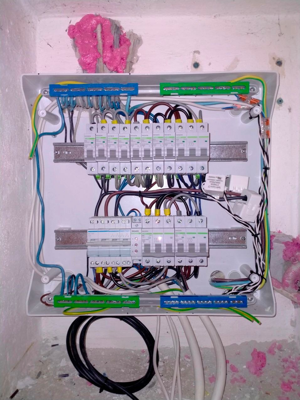 Otwarta skrzynka elektryczna z zamontowanymi bezpiecznikami i przewodami, widoczne ślady pianki montażowej na ścianie.