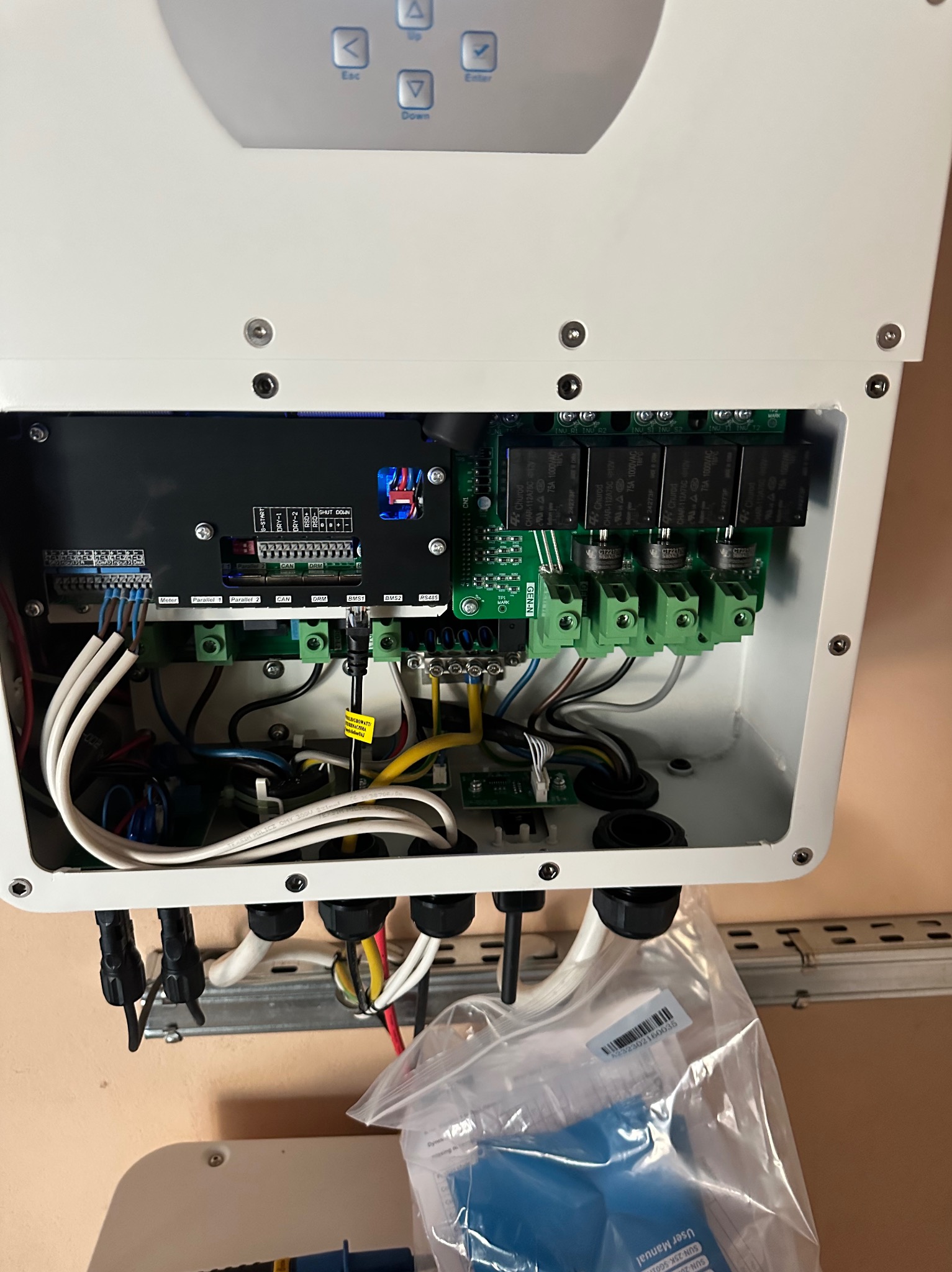 Wnętrze białej obudowy falownika z widocznymi przewodami, złączami, płytkami drukowanymi i elementami elektronicznymi. W dolnej części widoczna torba z instrukcją obsługi falownika.