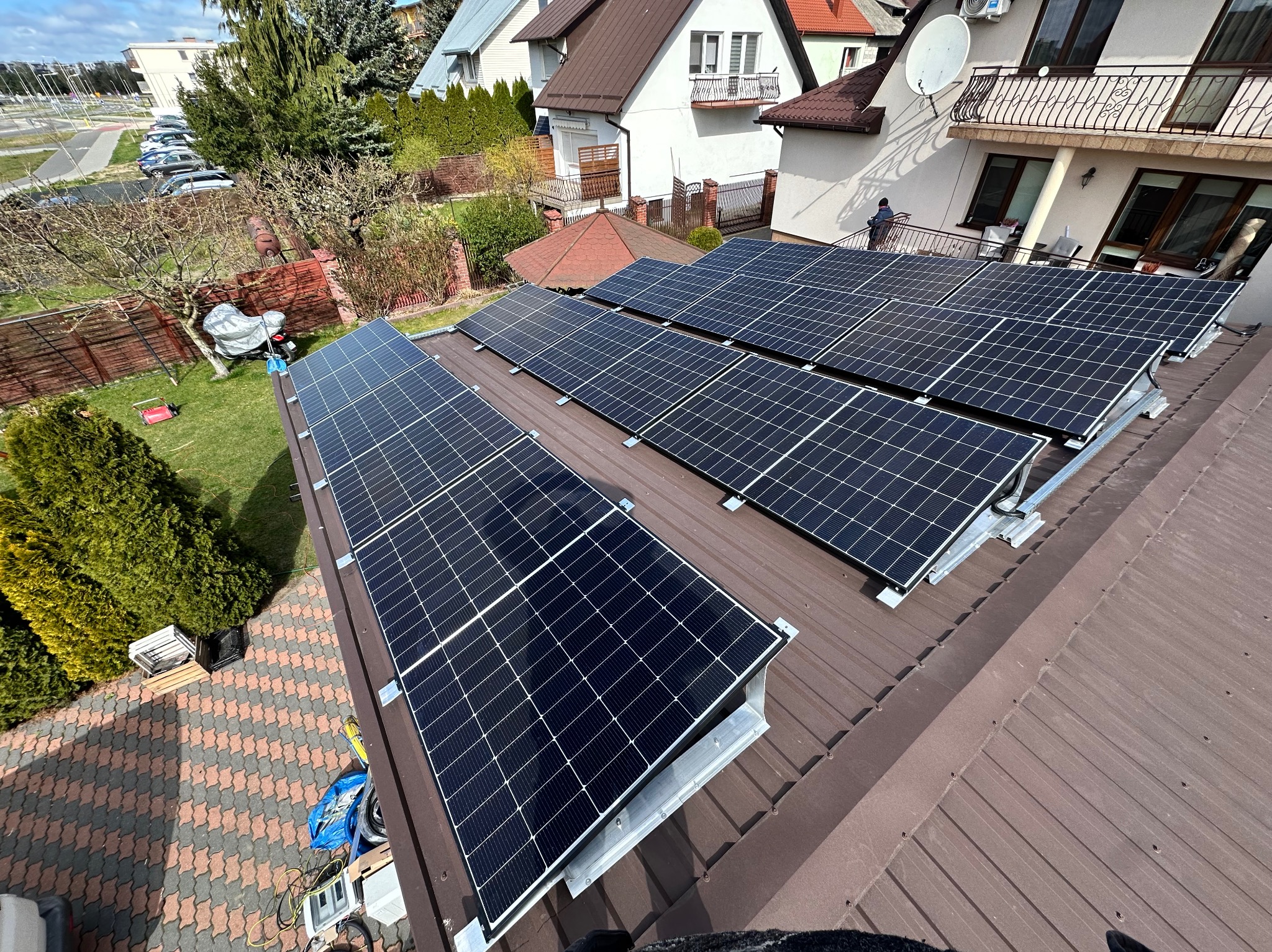 Rząd paneli słonecznych na brązowym dachu domu jednorodzinnego, widok z góry, w tle zabudowa mieszkalna i zieleń.