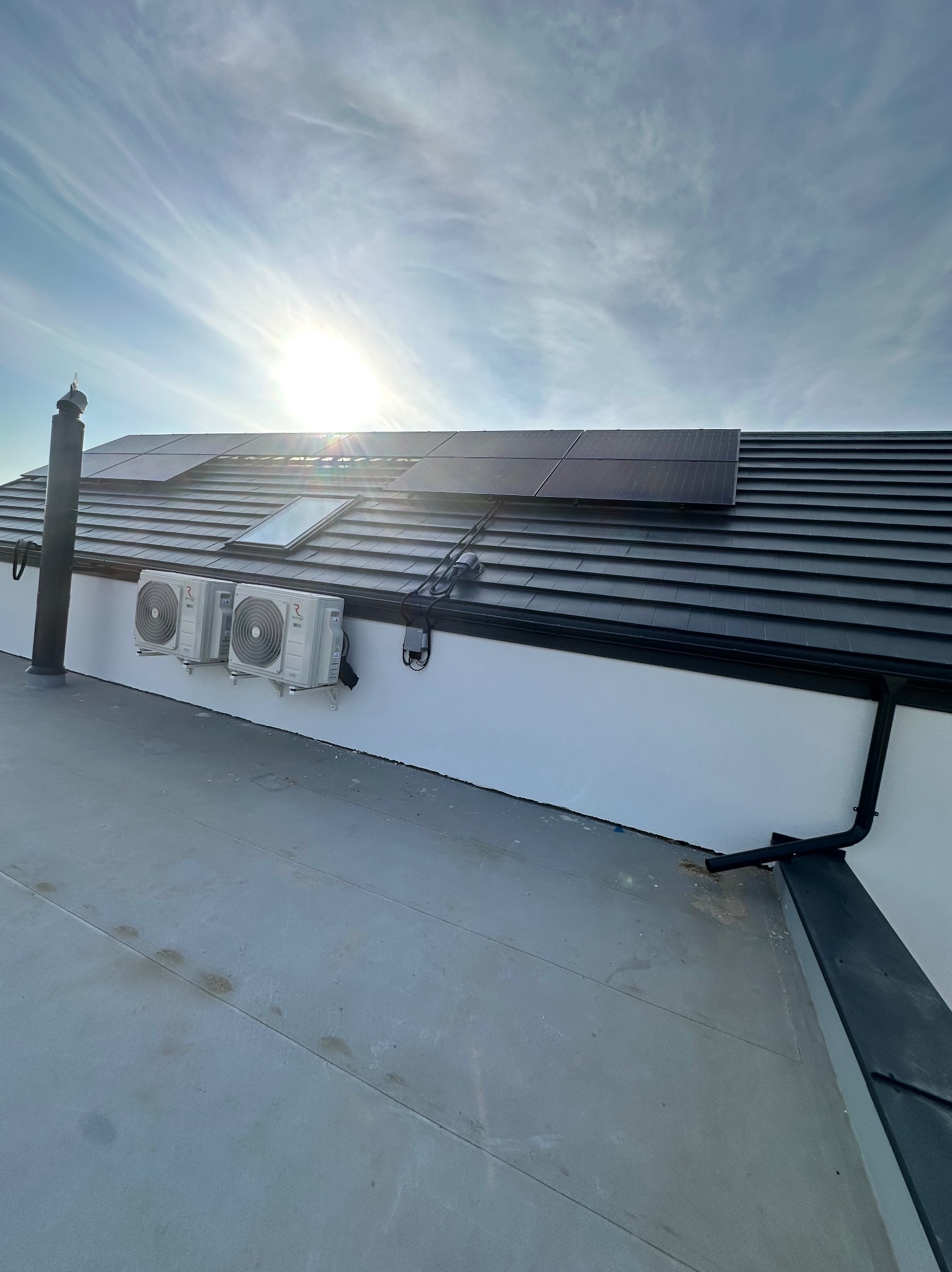 Panele słoneczne na dachu z dachówką, dwa klimatyzatory zamontowane na białej ścianie budynku, widoczne rury spustowe i okno dachowe, jasne słońce na niebie.