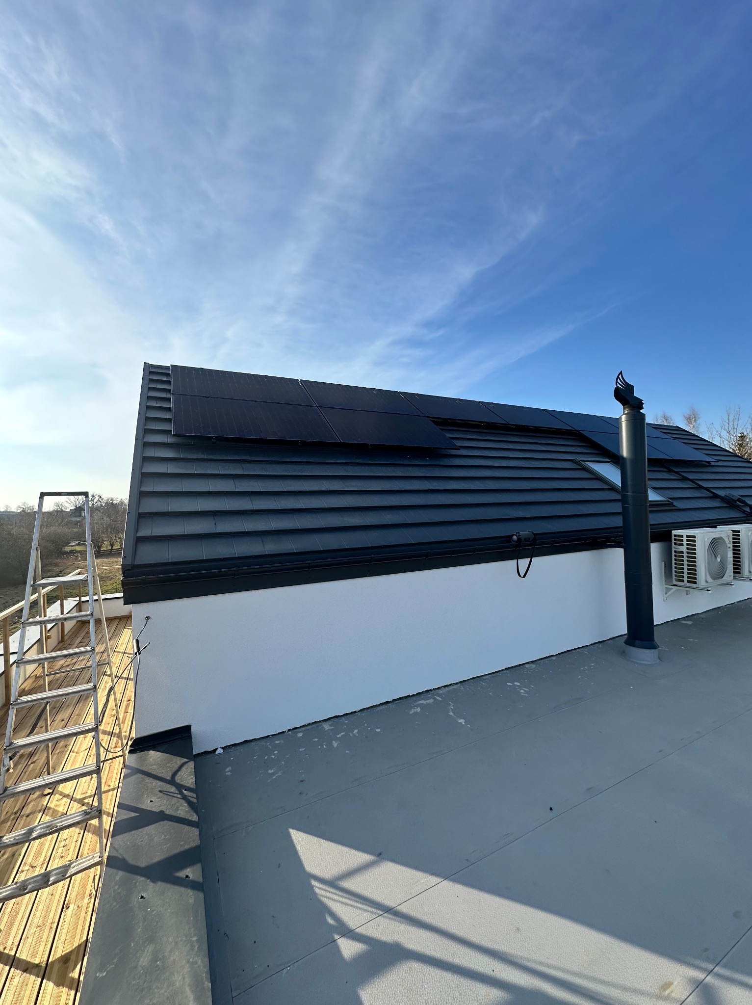 Czarny dach z panelami fotowoltaicznymi i kominem wentylacyjnym w słoneczny dzień, widoczne dwa klimatyzatory na białej ścianie oraz drabina na drewnianym tarasie.