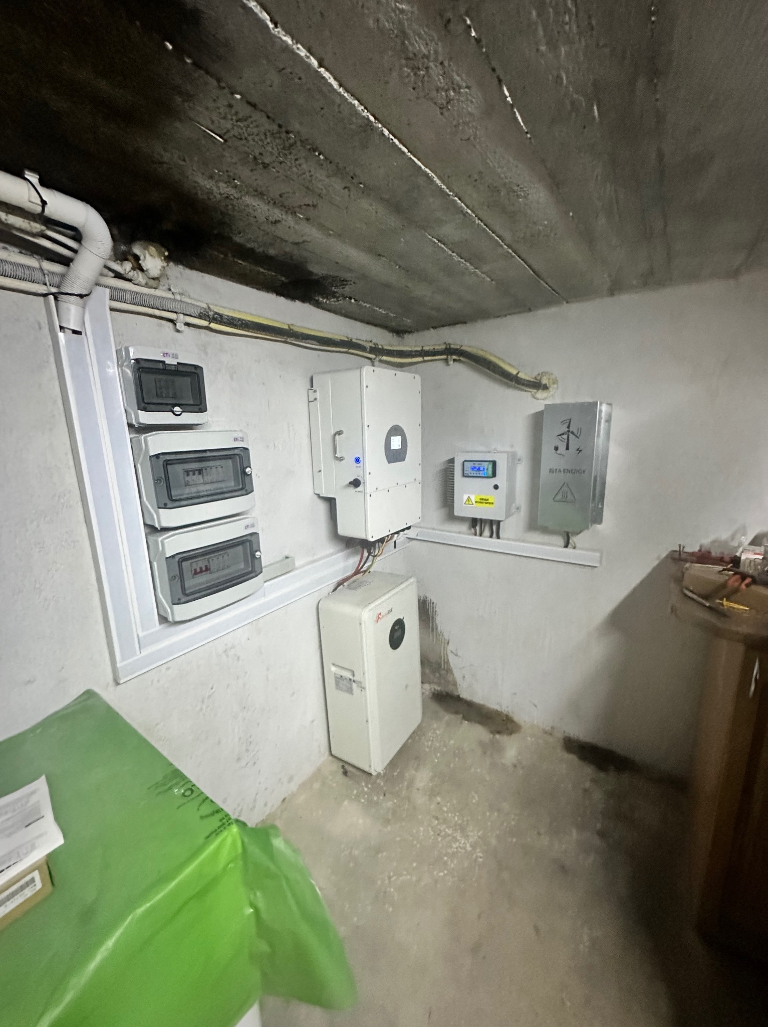 Instalacja urządzeń w pomieszczeniu piwnicznym: falownik, licznik energii, rozdzielnica, akumulator, okablowanie. Widoczny surowy betonowy sufit i ślady wilgoci na ścianach.