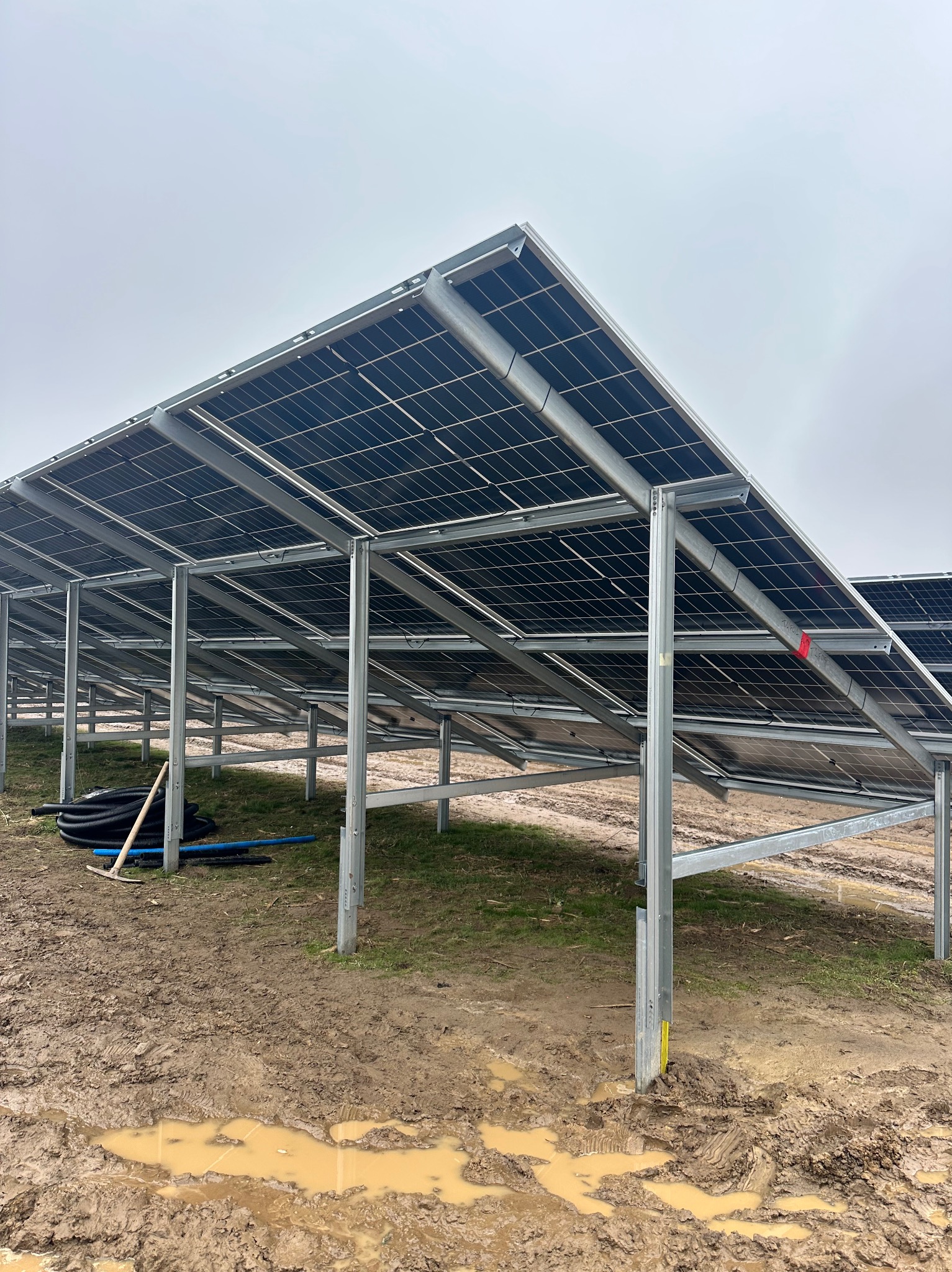 Rząd paneli słonecznych w trakcie montażu, widok od spodu, z widoczną konstrukcją wsporczą i przewodami na błotnistym terenie w pochmurny dzień.