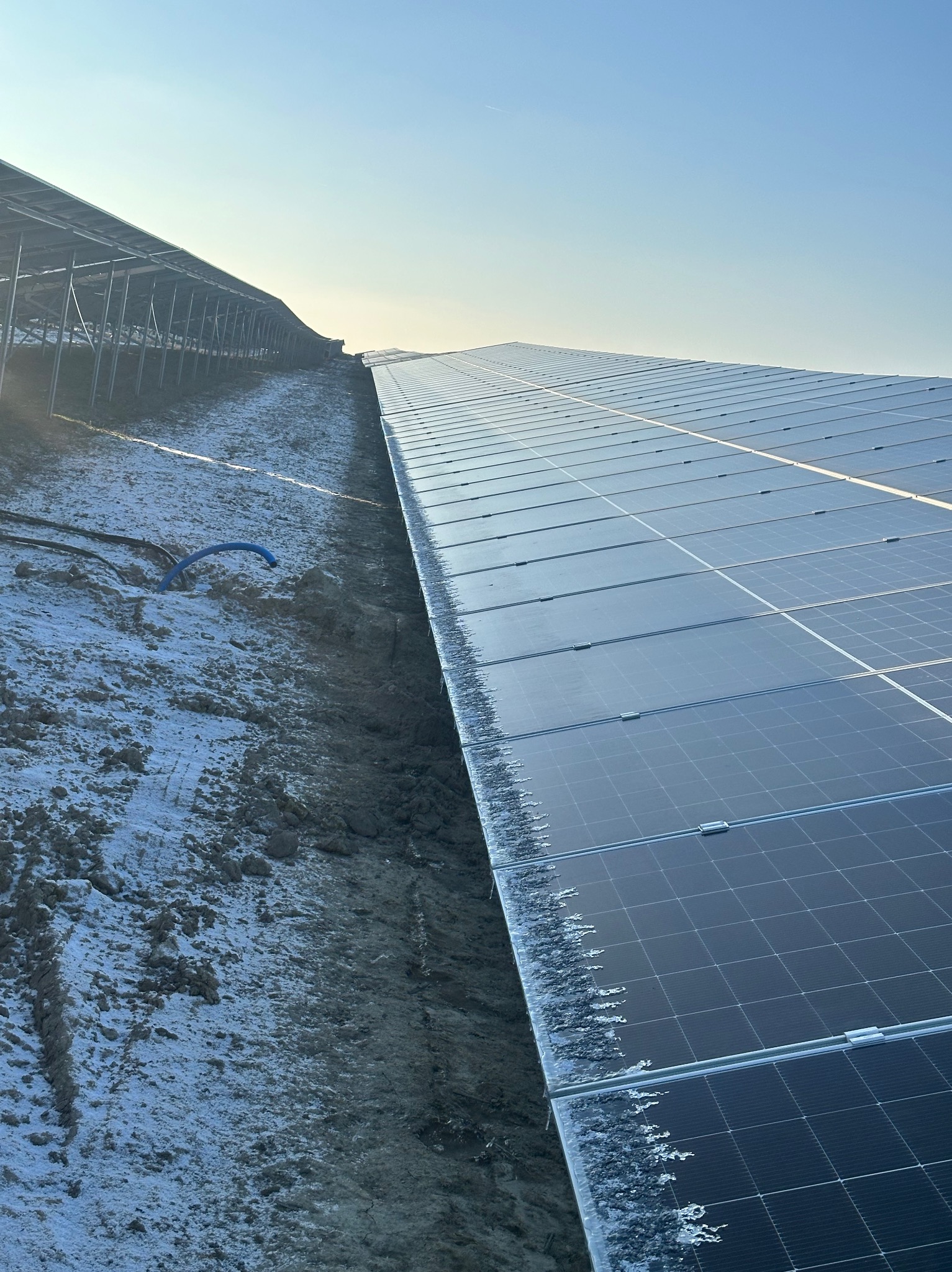 Długi rząd paneli słonecznych pokrytych szronem, zamontowanych na ziemi, widoczny błękitny przewód i metalowa konstrukcja w tle, pochmurne niebo.