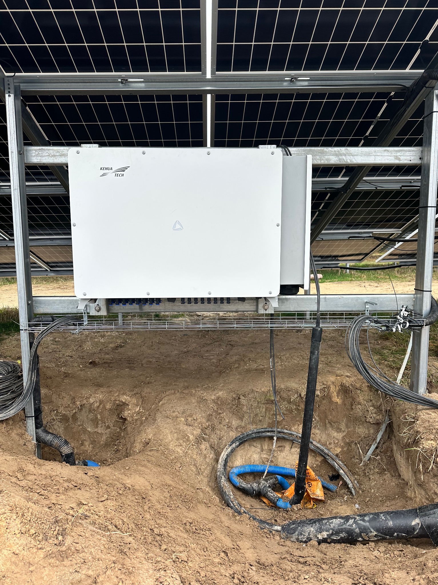 Montaż instalacji z panelami słonecznymi: widoczny biały inwerter marki Kehua Tech przymocowany do konstrukcji pod panelami, z przewodami w ziemi w wykopie, niebieska rura karbowana i pomarańczowa...