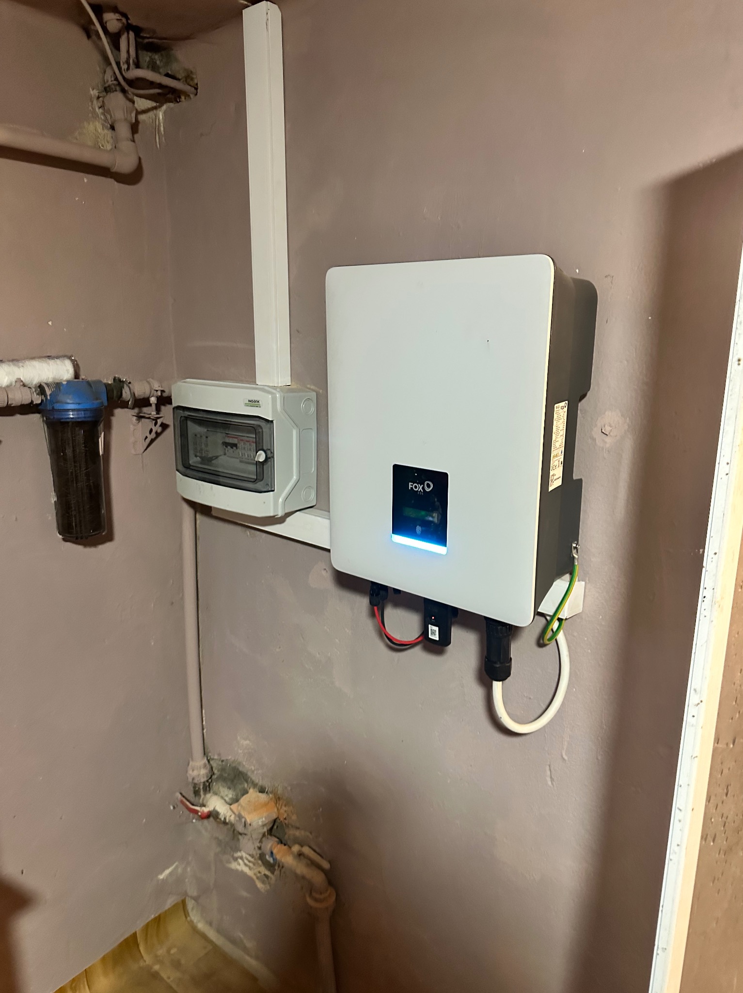 Biały inwerter fotowoltaiczny marki FOX zamontowany na ścianie w pomieszczeniu technicznym, widoczna skrzynka z bezpiecznikami i filtr na rurze.