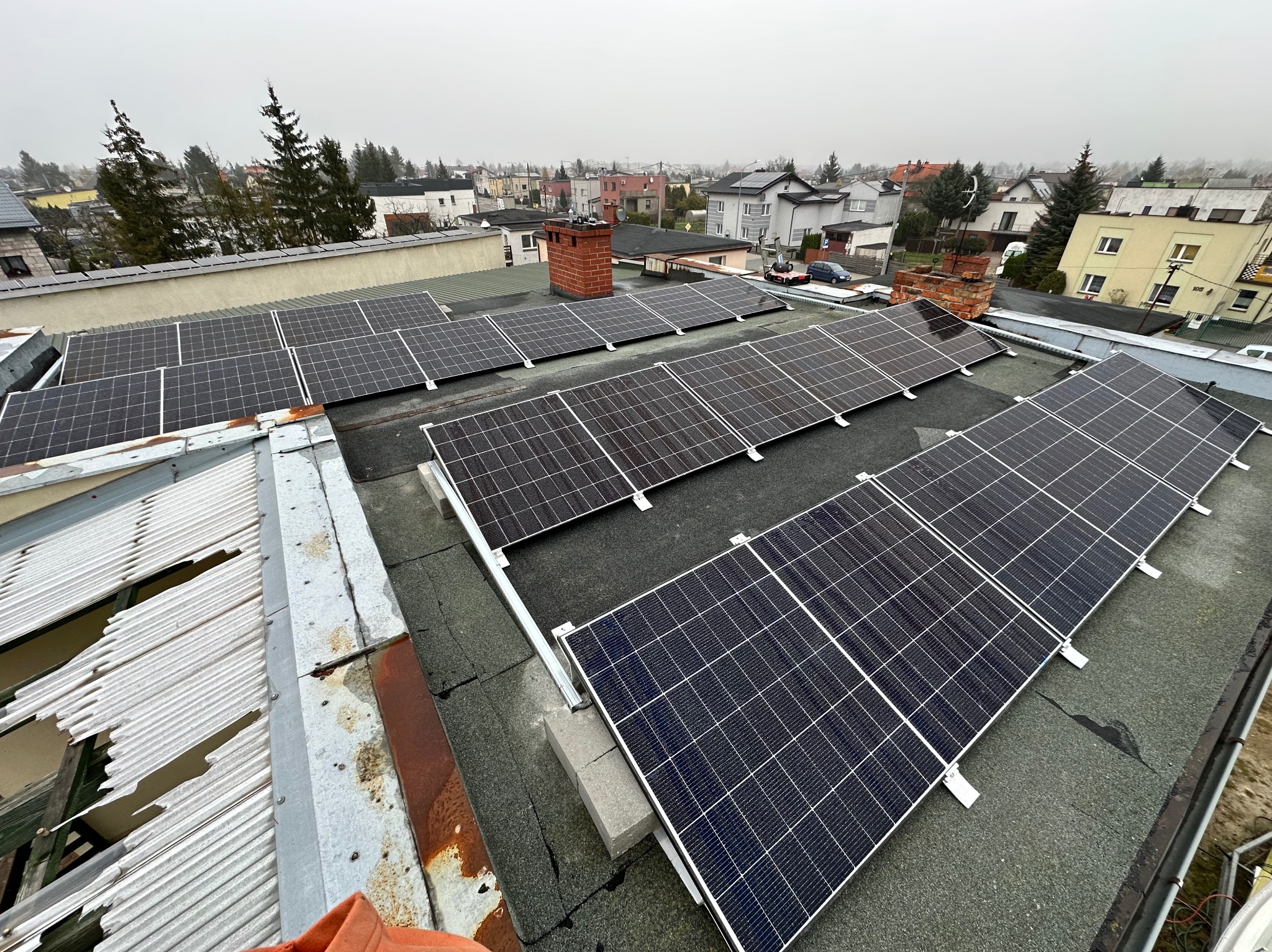 Widok z góry na instalację paneli słonecznych na płaskim dachu w Gdańsku, z widoczną okoliczną zabudową miejską w pochmurny dzień.