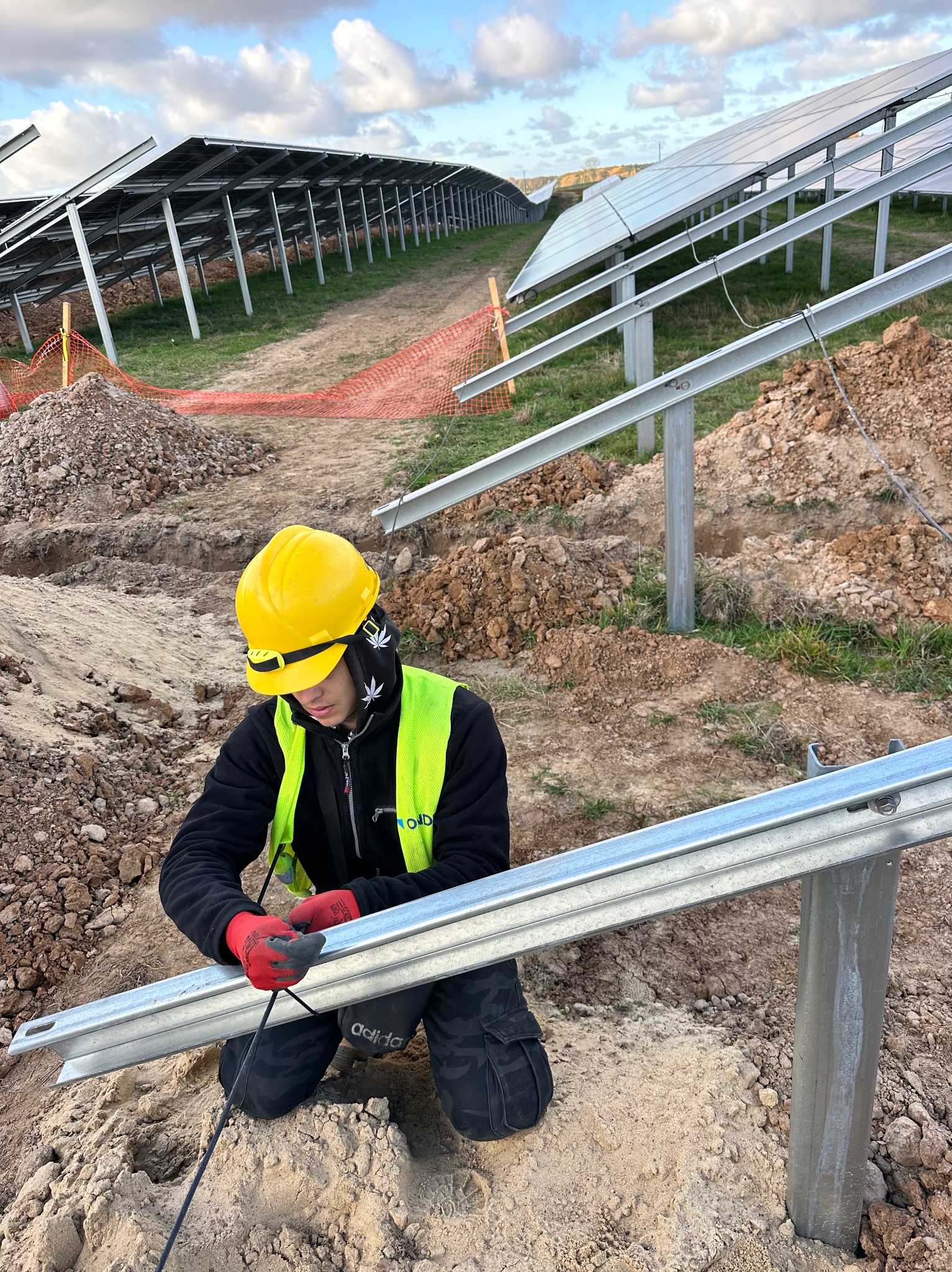 Pracownik w kasku i kamizelce odblaskowej, w czerwonych rękawicach, przywiązuje metalową konstrukcję pod panele słoneczne na tle częściowo zamontowanej instalacji na gruncie.