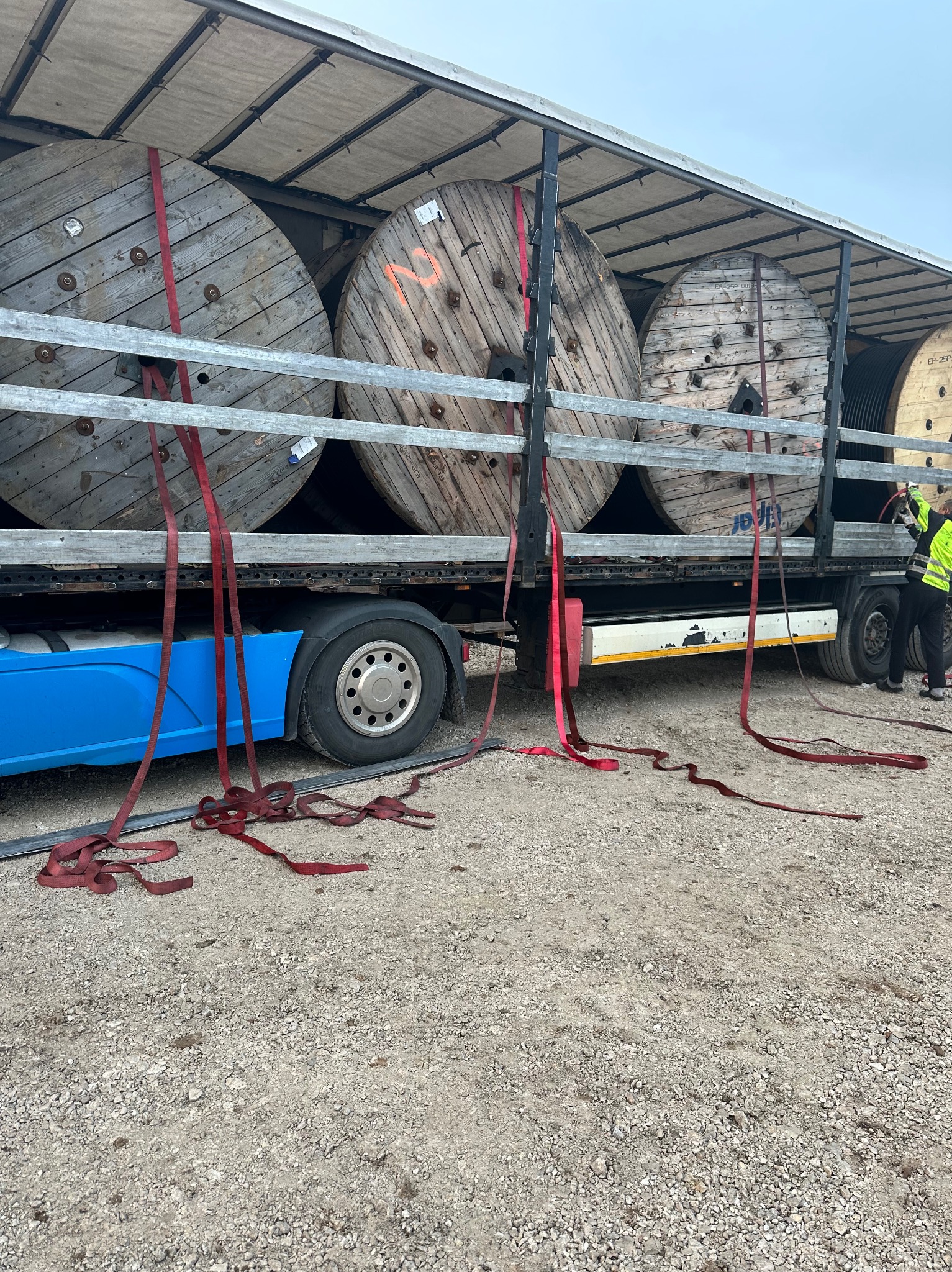 Transport kabli na drewnianych bębnach, zabezpieczonych czerwonymi pasami, na platformie ciężarówki z niebieską kabiną, widok z boku, na żwirowym podłożu.