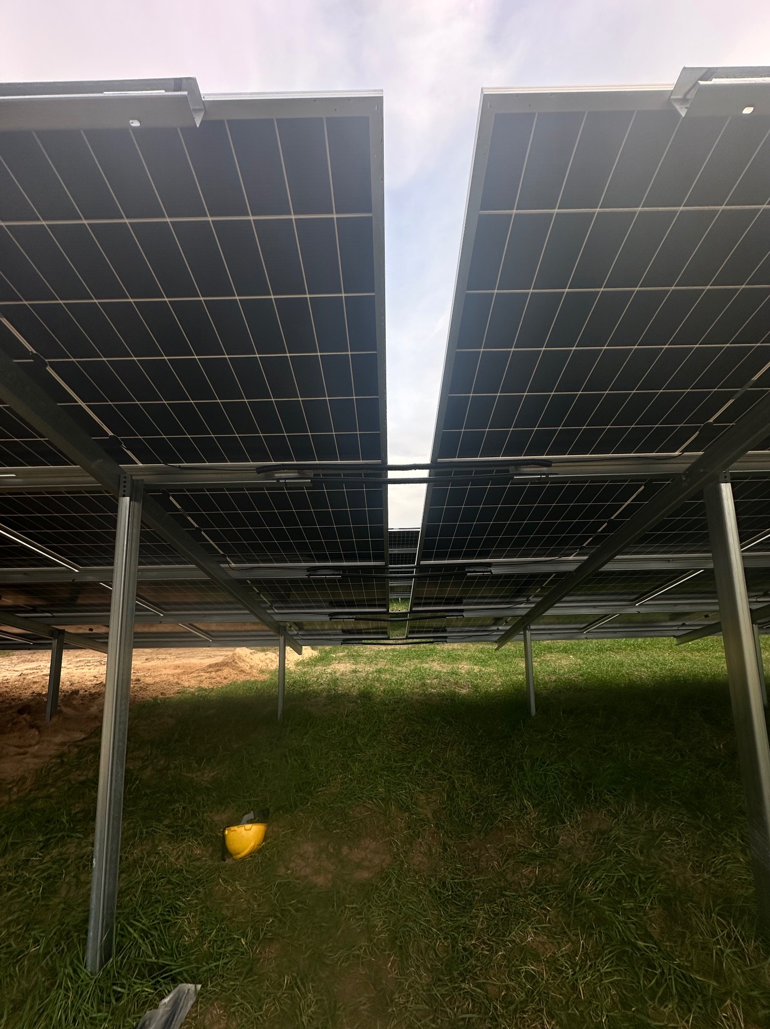 Widok z dołu na konstrukcję wsporczą paneli słonecznych na trawie, z żółtym kaskiem leżącym obok jednej z nóg wspornika.