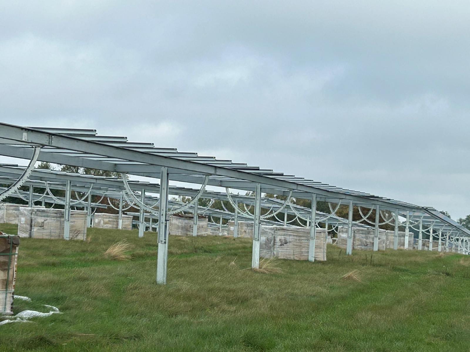 Stalowa konstrukcja wsporcza pod panele, przygotowana do montażu na trawiastym terenie, pakiety paneli zabezpieczone folią czekają w tle.