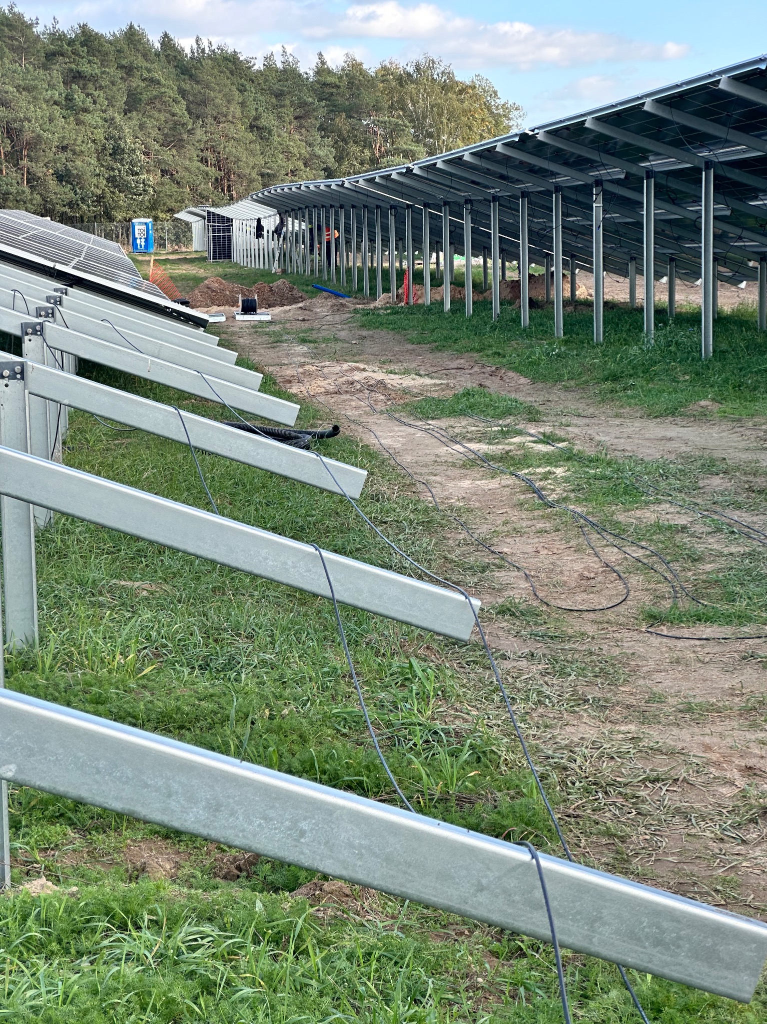 Montaż konstrukcji wsporczej dla paneli słonecznych na terenie zielonym, widoczne kable i tymczasowa toaleta TOI TOI w tle.
