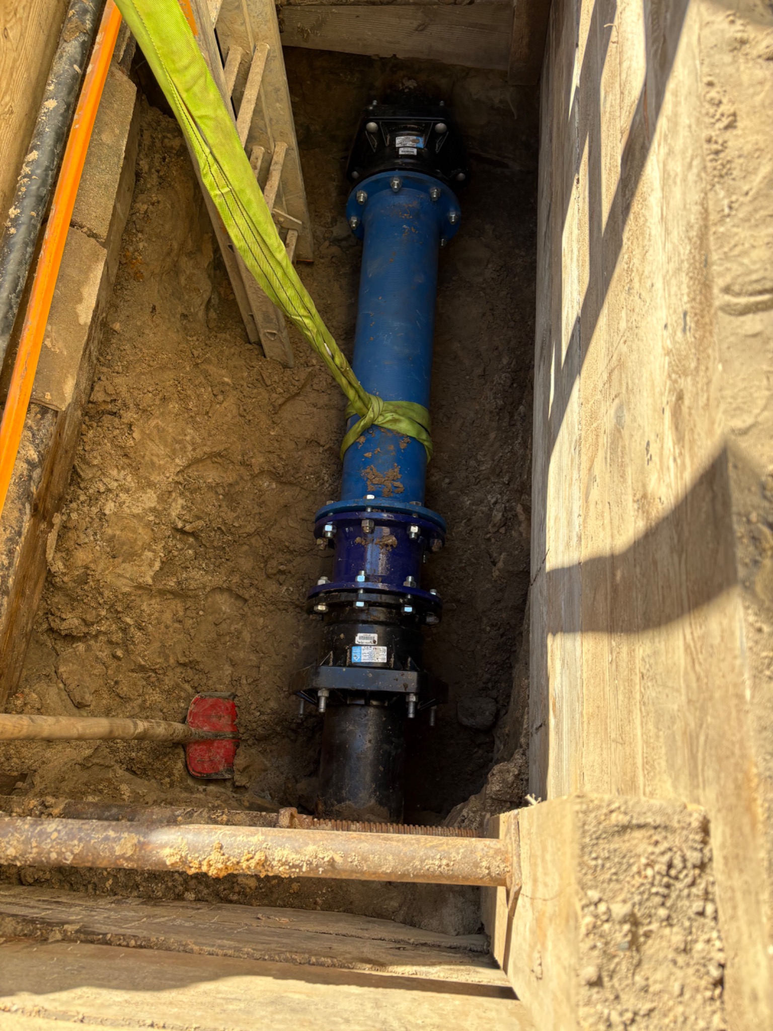 Niebieska rura wodociągowa z kołnierzami, zabezpieczona pasem transportowym, umieszczona w wykopie z drewnianą obudową, widoczna łopata i drabina.