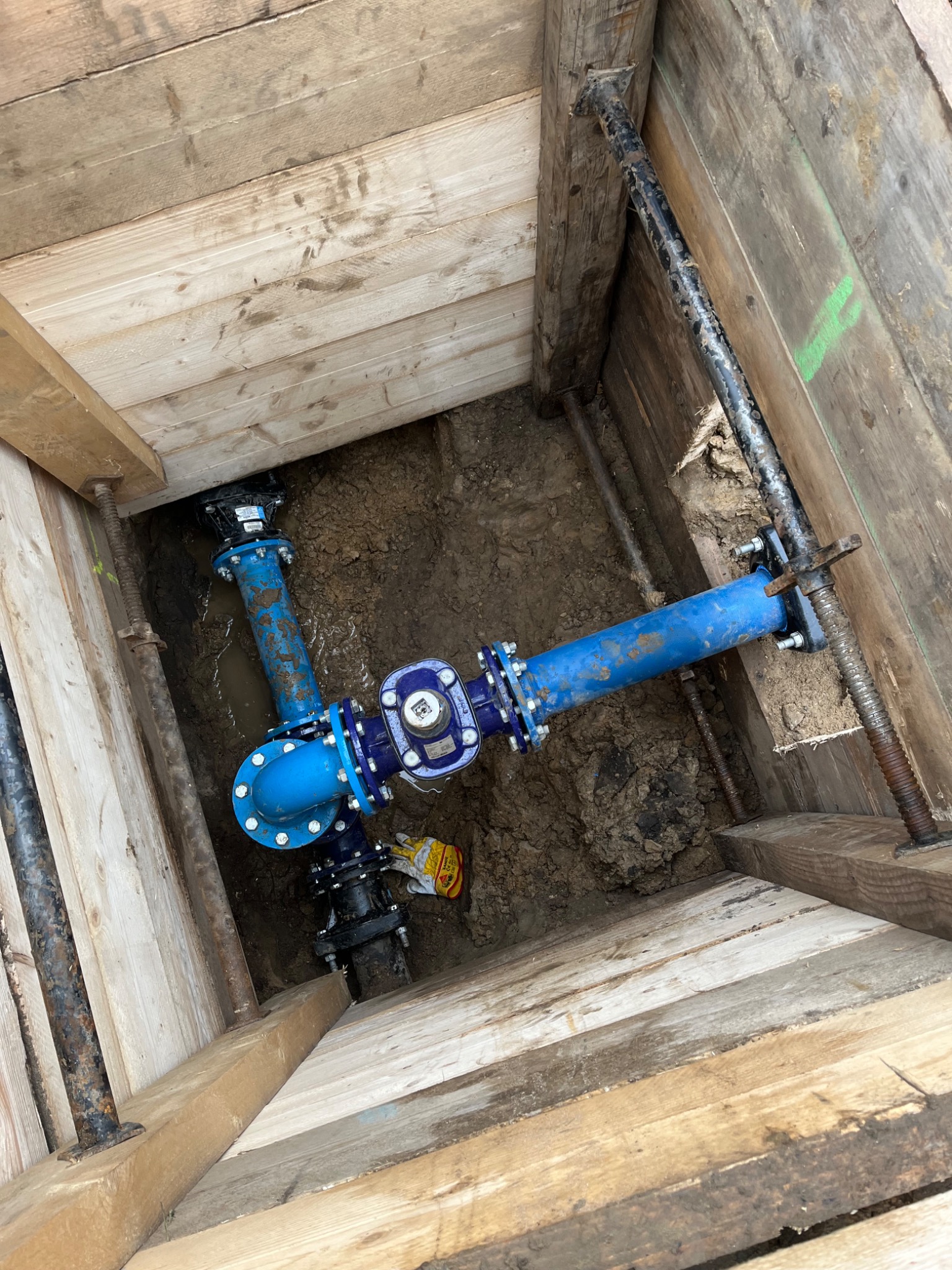 Wykonane przyłącze wodociągowe w wykopie zabezpieczonym drewnianą szalunką, widoczne niebieskie rury i złączki, elementy mocujące oraz fragment żółtej rękawicy roboczej.
