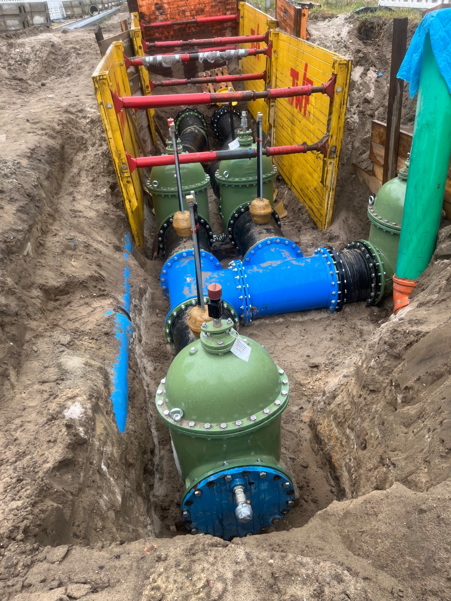 Wykop z zabezpieczeniami ścian, widoczne rury wodociągowe i zawory w kolorach zielonym i niebieskim, częściowo zasypane ziemią.