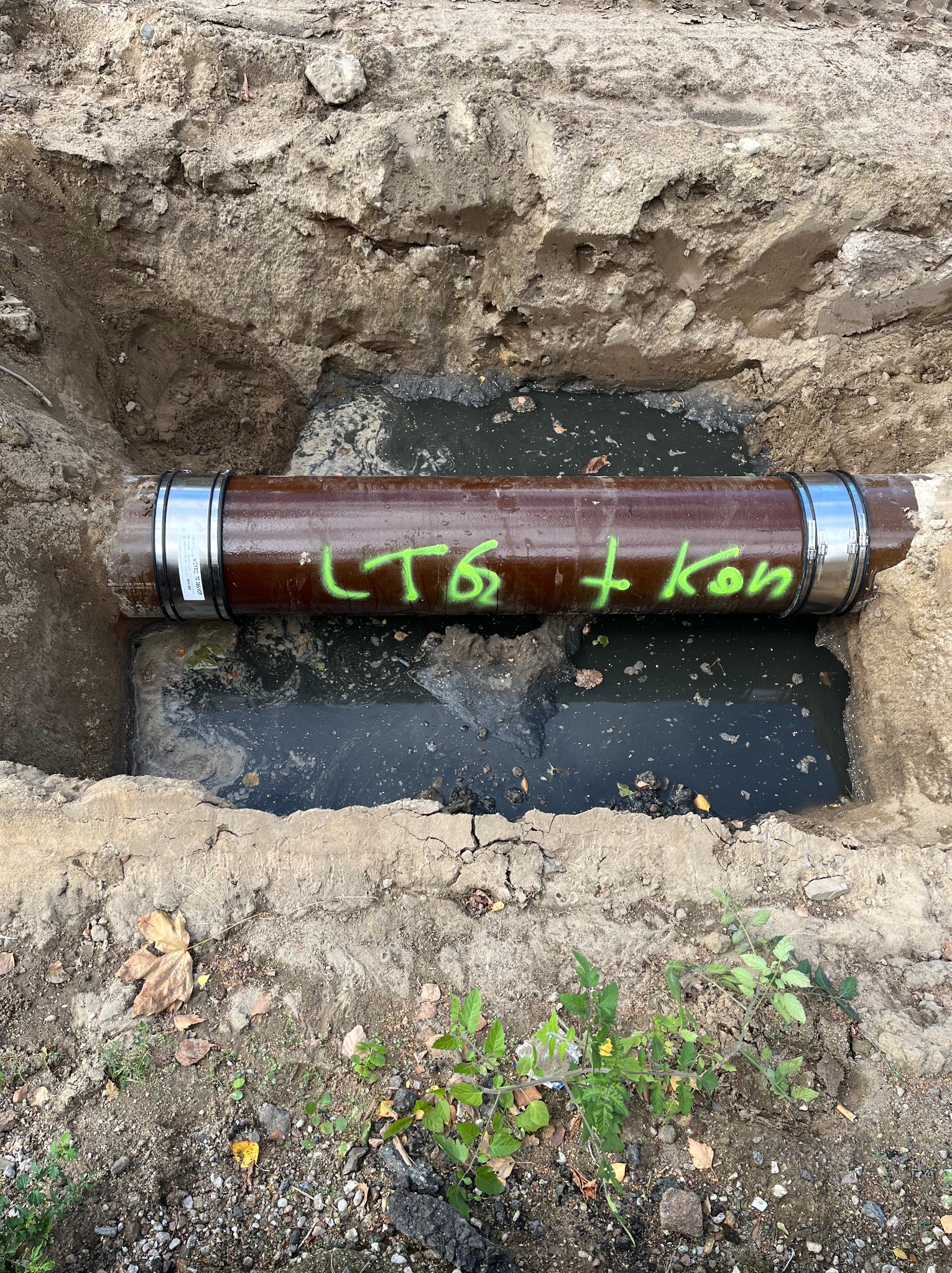 Brązowa rura z metalowymi obejmami leżąca w wykopie wypełnionym wodą, na rurze zielony napis 'LTG + kon'