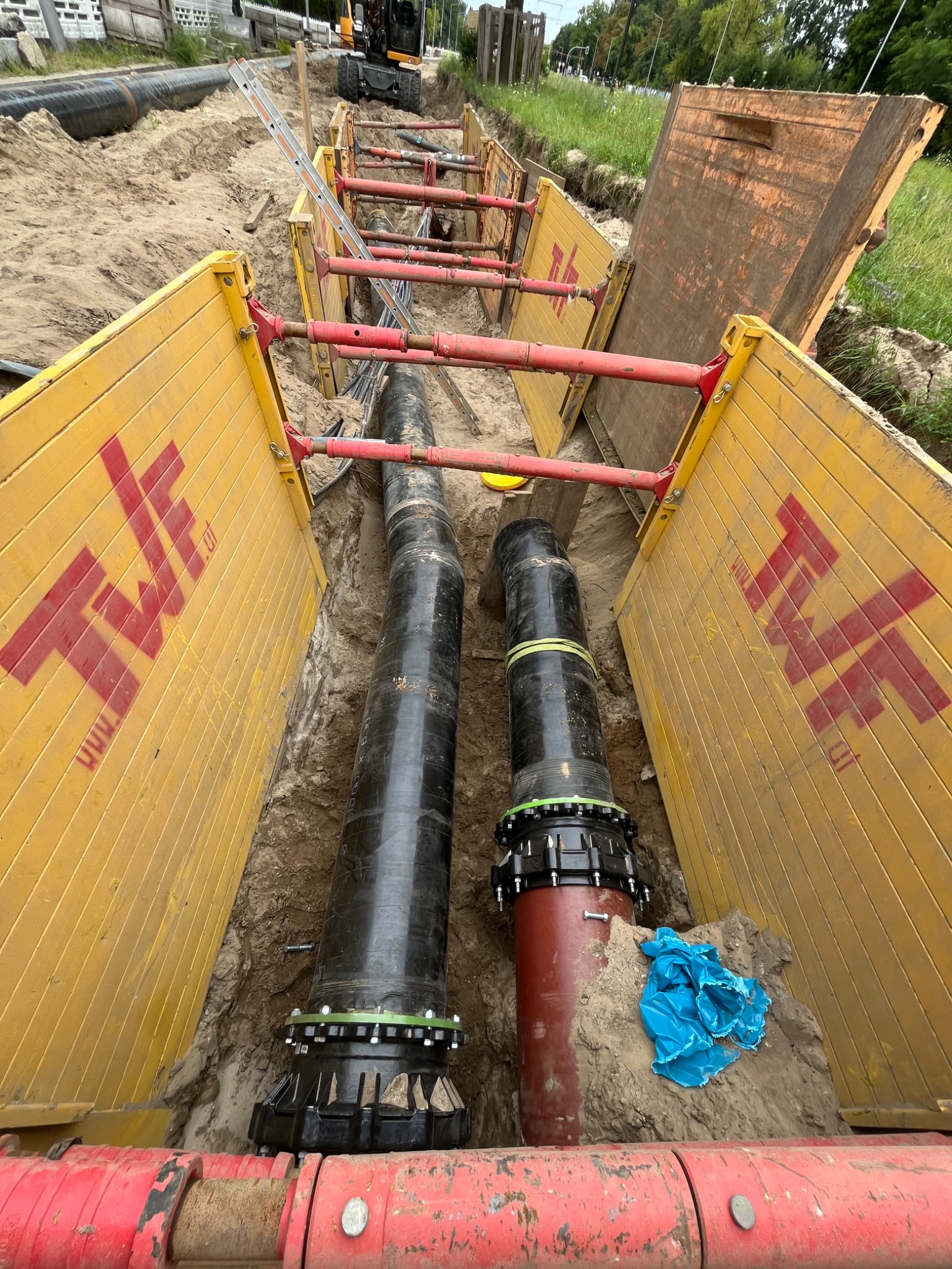 Wykop zabezpieczony żółtymi panelami z widocznymi rurami kanalizacyjnymi w trakcie instalacji, na tle widoczny sprzęt budowlany i zieleń.