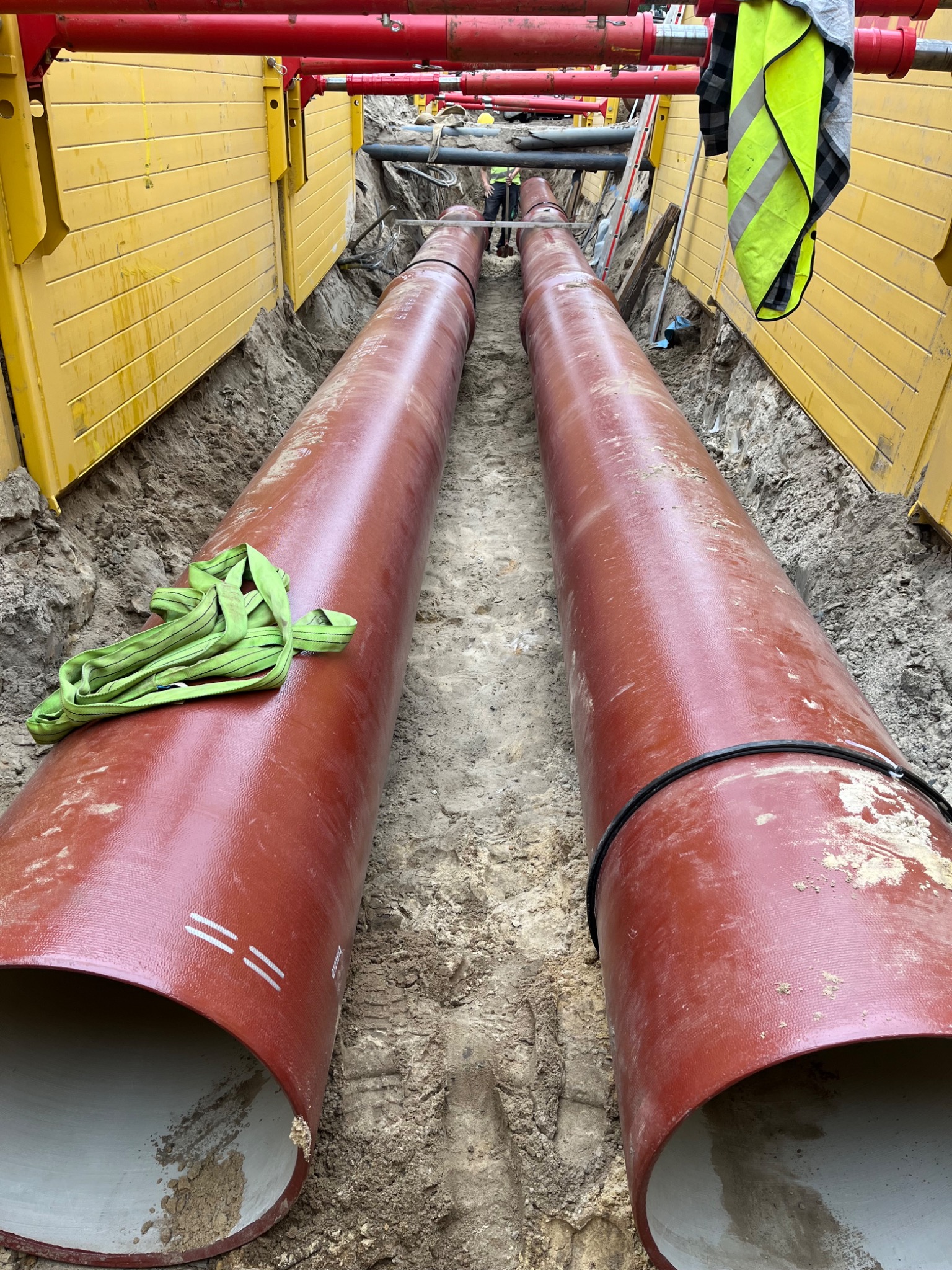 Dwa długie, brązowe rury kanalizacyjne ułożone w wykopie zabezpieczonym żółtymi panelami, z widocznymi elementami zabezpieczającymi wykop i zielonym pasem transportowym na jednej z rur.