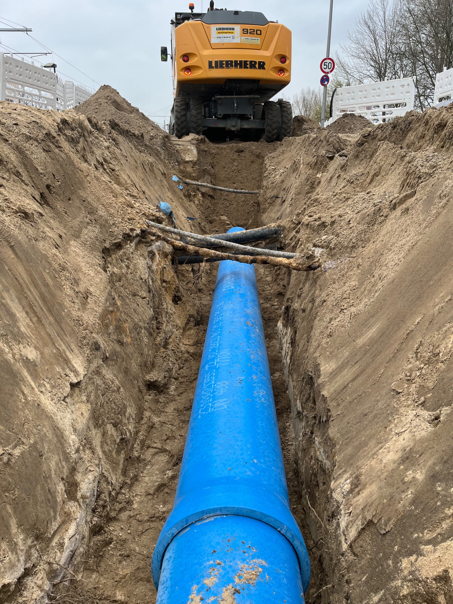 Głęboki wykop z ułożoną, niebieską rurą wodociągową, widoczna koparka Liebherr 920 i znak ograniczenia prędkości w tle, ziemia odsłania warstwy geologiczne.