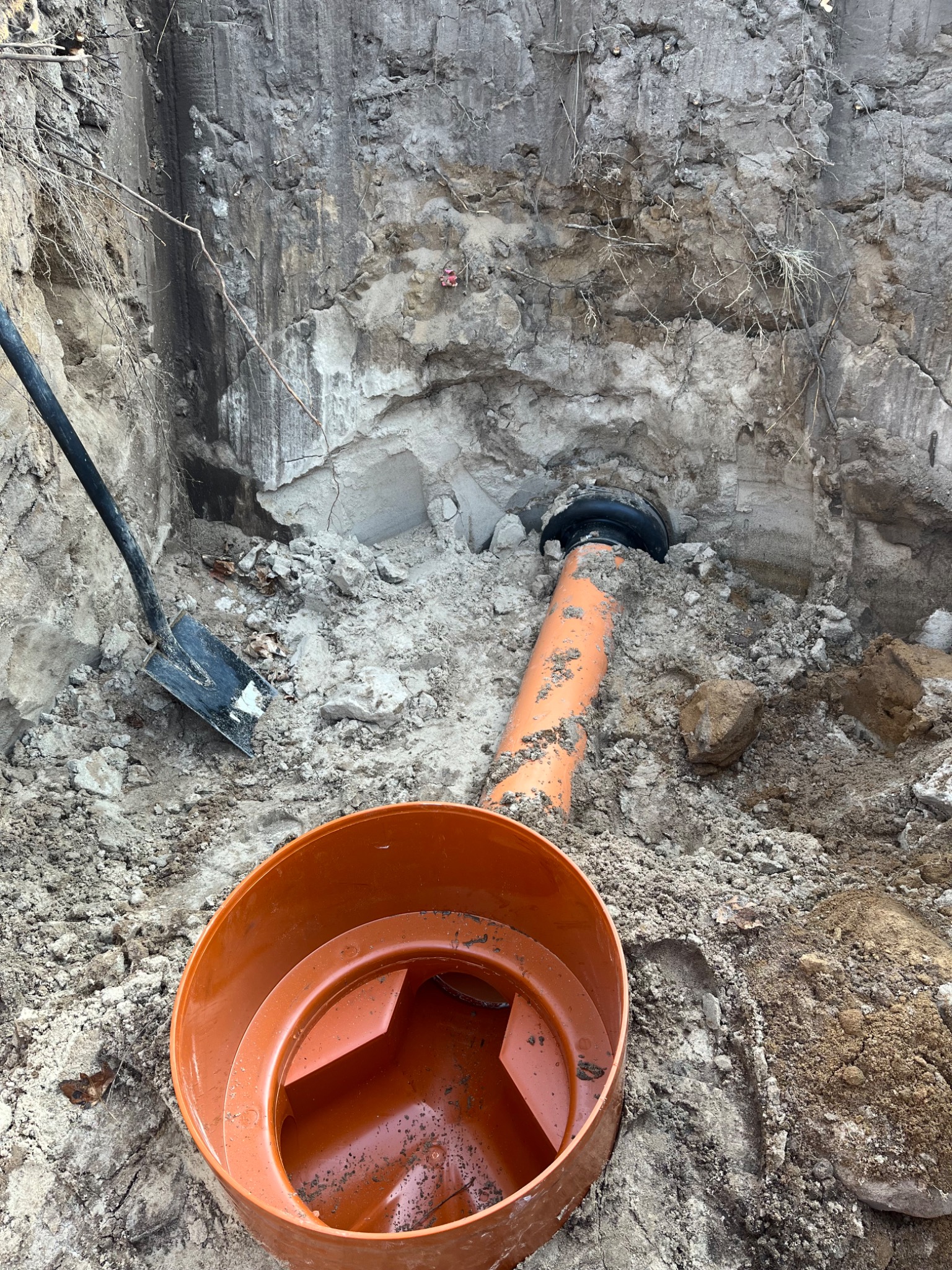 Pomarańczowa rura kanalizacyjna w wykopie ziemnym, czarna złączka, widoczna łopata i element szamba betonowego.