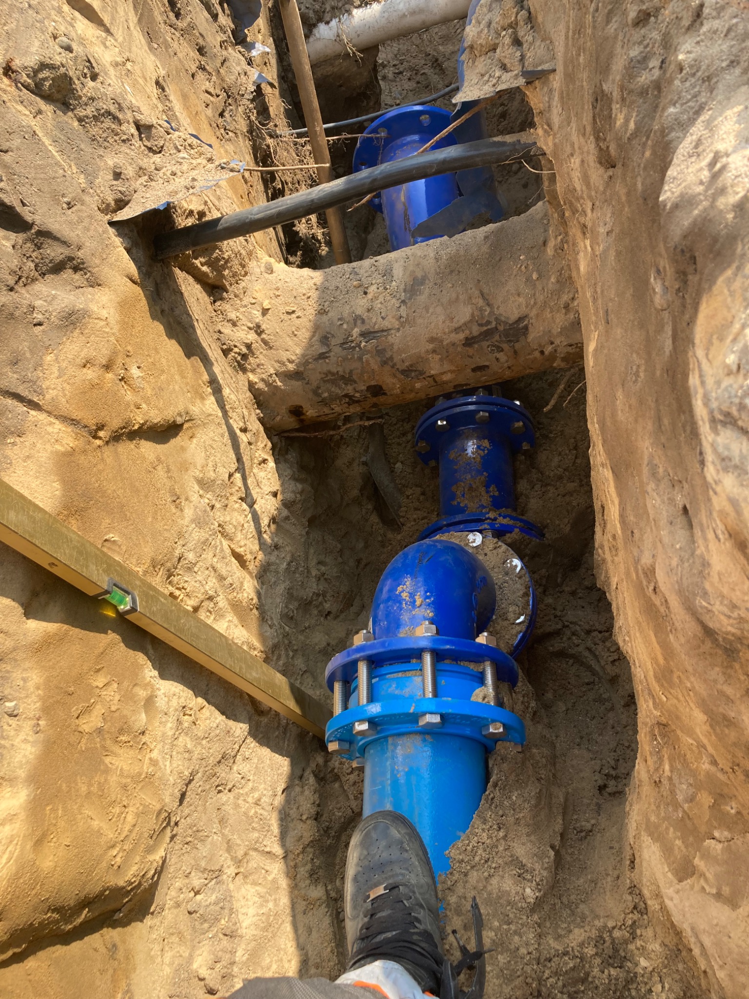 Niebieska rura wodociągowa z kołnierzami w wykopie ziemnym, poziomica przyłożona do ściany wykopu, widoczny fragment buta pracownika.