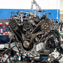 SV - Motors - Silnik samochodowy zawieszony na łańcuchu w warsztacie, widoczne koła zębate i łańcuch rozrządu. Na tle ściana warsztatu z narzędziami.