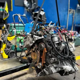 Wyjęty silnik samochodowy zawieszony na pasach w warsztacie, widoczne elementy mechaniczne i przewody. Niebieska ściana w tle.