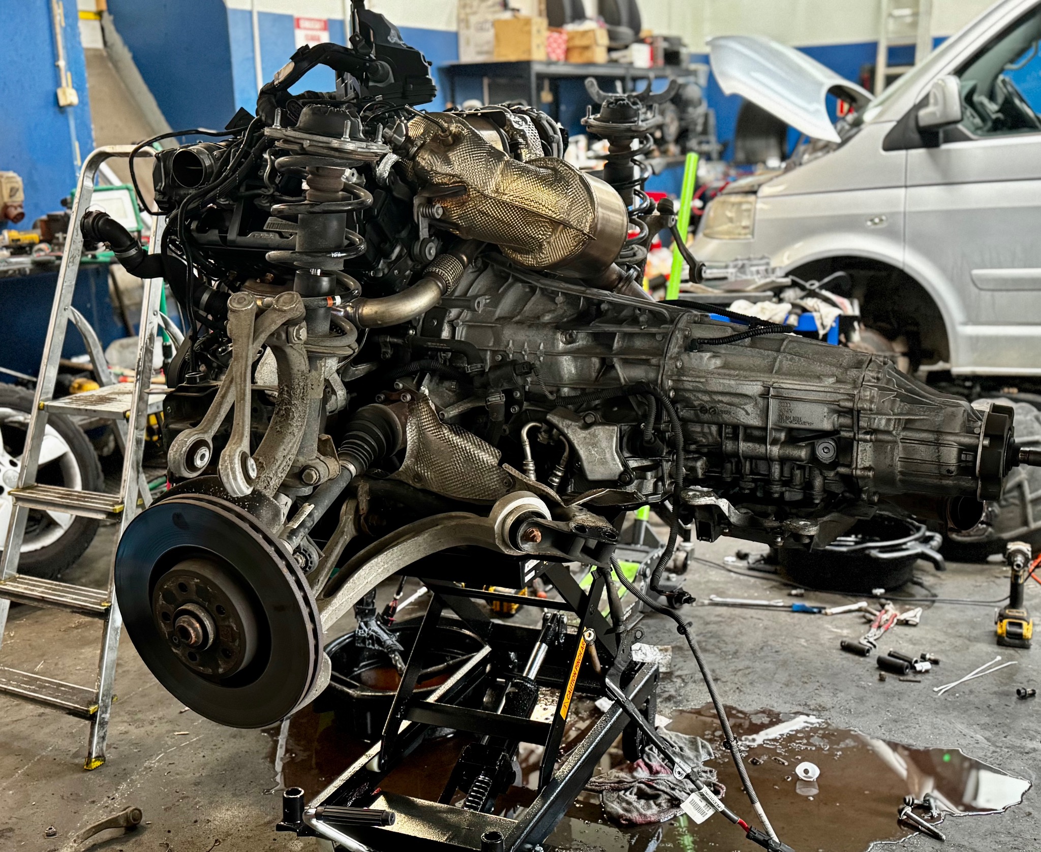 Wyjęty silnik samochodowy na podnośniku w warsztacie. Widoczne elementy zawieszenia, tarcza hamulcowa, skrzynia biegów. W tle samochód z otwartą maską i narzędzia.