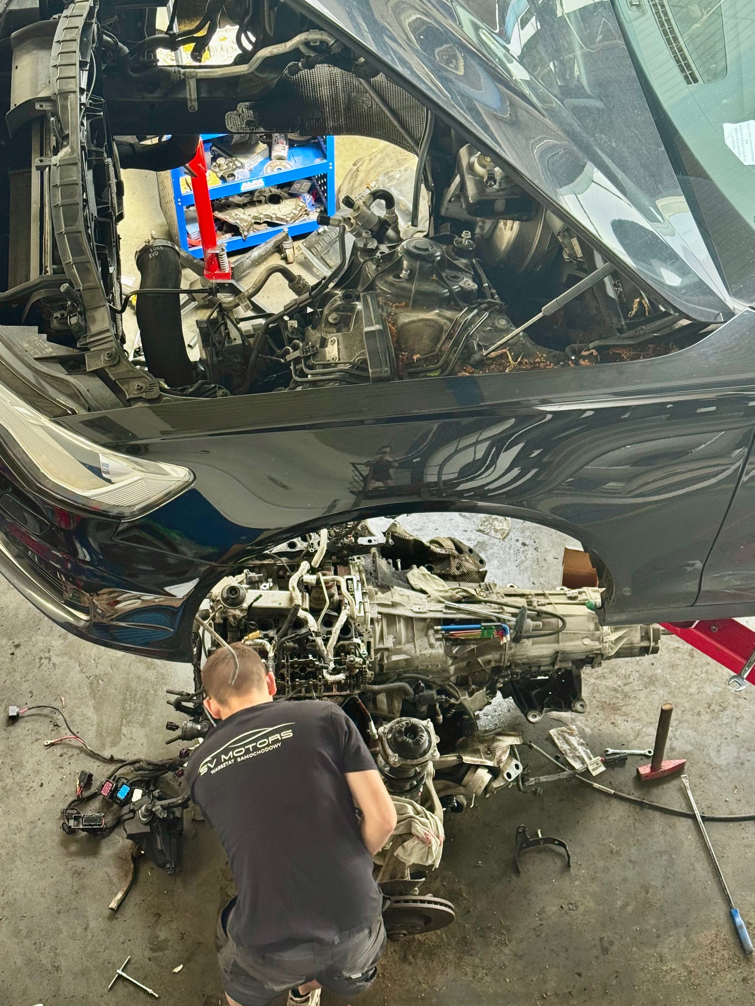 Czarny samochód z otwartą maską, silnik wyjęty z auta, mechanik w koszulce SV Motors przy pracy. W tle widać narzędzia i wyposażenie warsztatu.