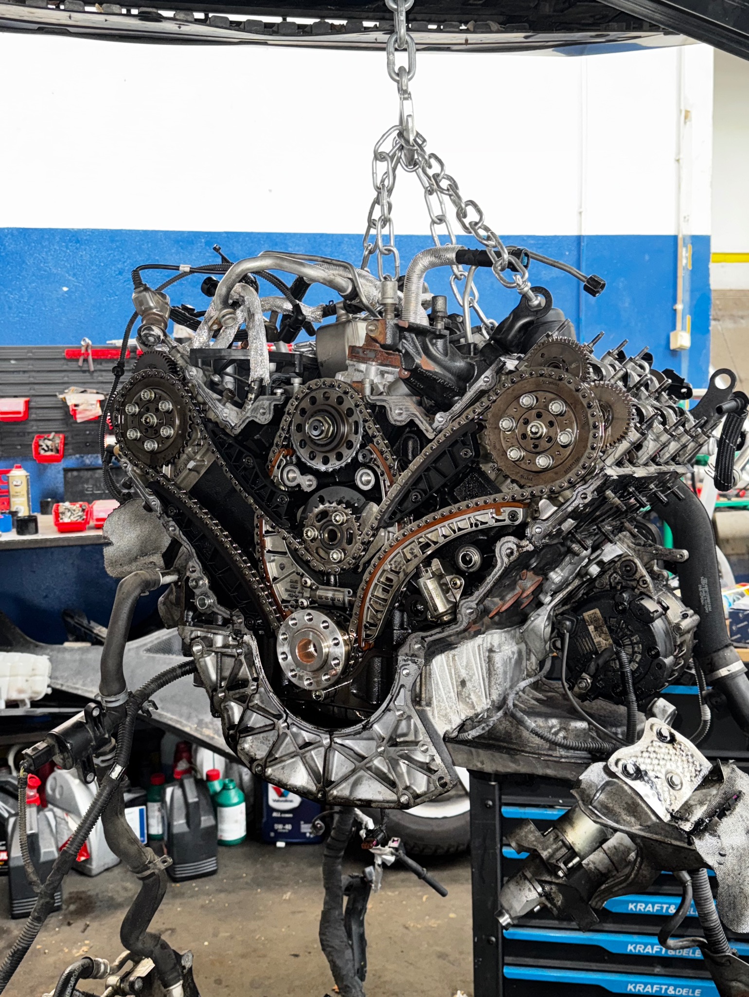 Silnik samochodowy zawieszony na łańcuchu w warsztacie, widoczne koła zębate i łańcuch rozrządu. Na tle ściana warsztatu z narzędziami.