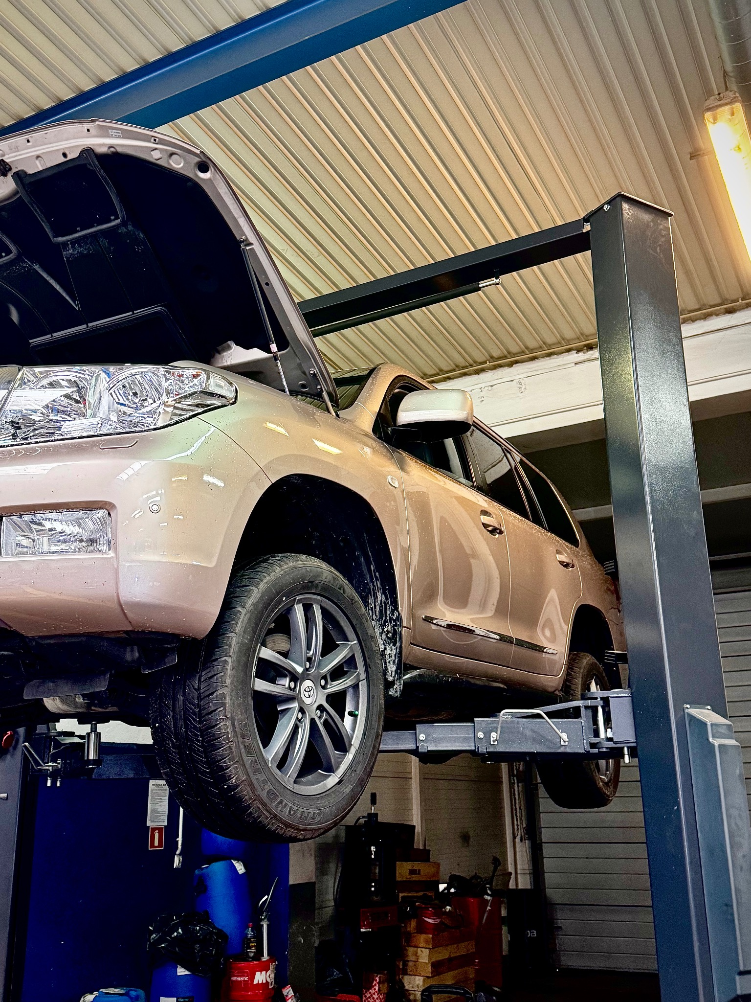 Beżowy SUV na podnośniku w warsztacie samochodowym, z otwartą maską. Widoczne detale oświetlenia i wyposażenia warsztatu.