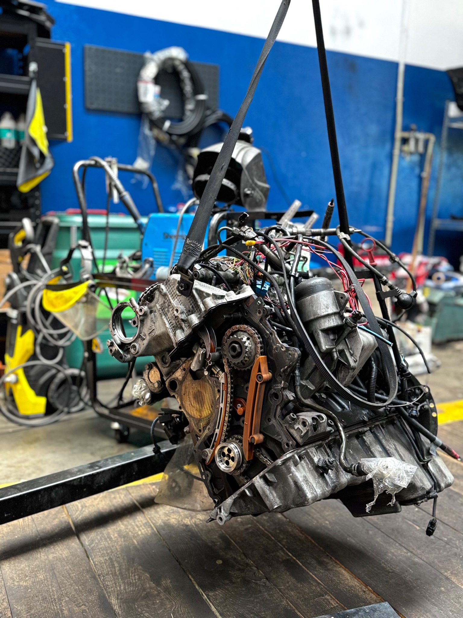 Wyjęty silnik samochodowy zawieszony na pasach w warsztacie, widoczne elementy mechaniczne i przewody. Niebieska ściana w tle.