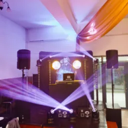 Scena z nagłośnieniem i oświetleniem na imprezie. Widoczne kolumny głośnikowe, reflektory i konsoleta DJ-a z grafiką muzyka w słuchawkach.