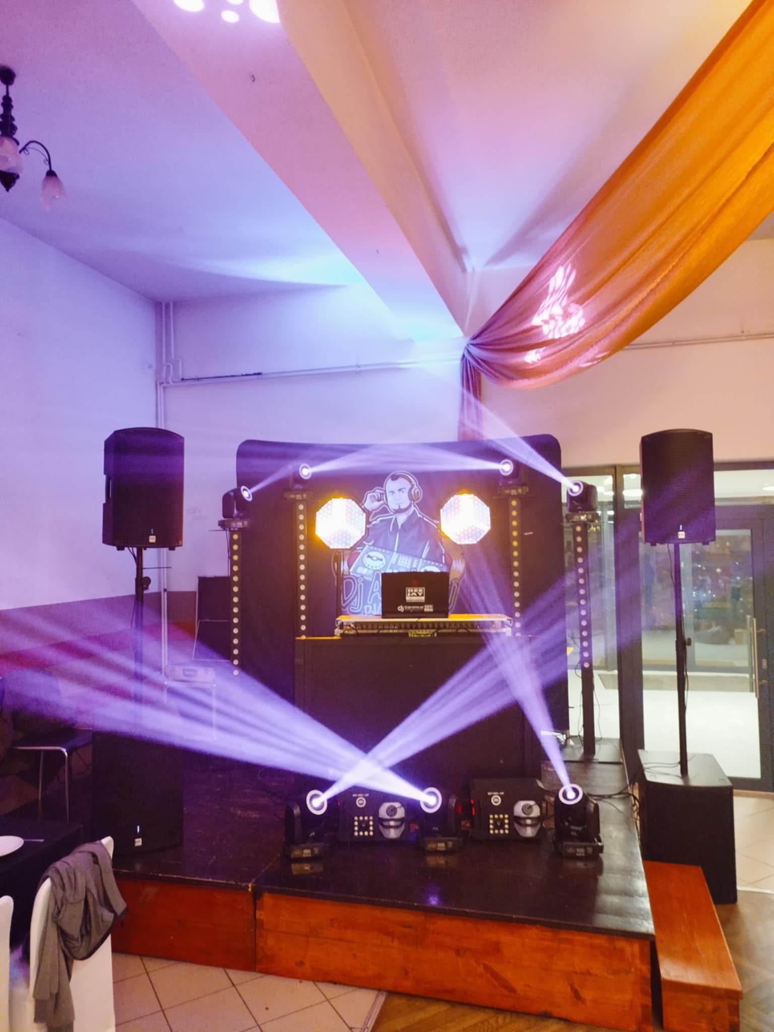 Scena z nagłośnieniem i oświetleniem na imprezie. Widoczne kolumny głośnikowe, reflektory i konsoleta DJ-a z grafiką muzyka w słuchawkach.