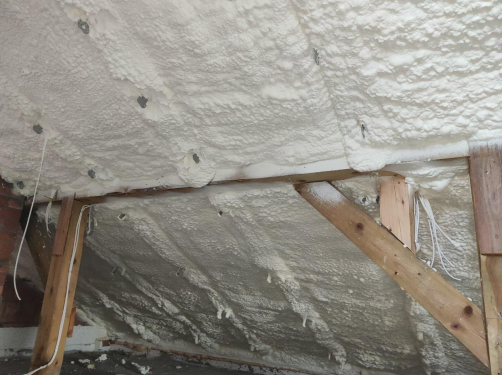 Poddasze ocieplone białą pianką poliuretanową, widoczne drewniane elementy konstrukcyjne oraz wystające przewody elektryczne.