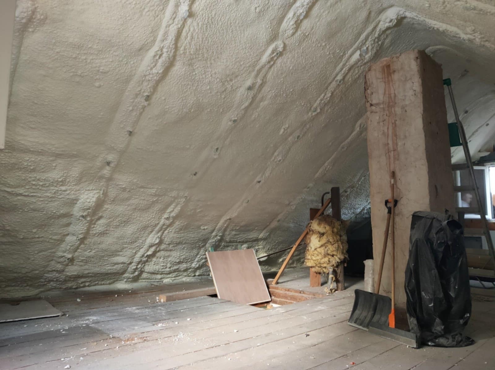 Poddasze po ociepleniu pianką PUR, widoczne nierównomierne pokrycie pianą, drewniana podłoga z resztkami materiałów budowlanych, oraz fragment kolumny i drabiny.