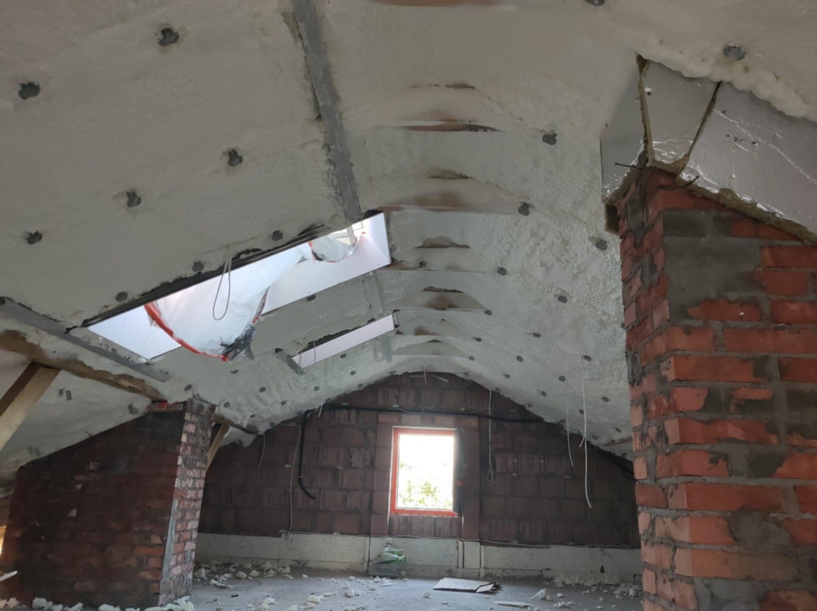Poddasze z ocieploną pianką PUR skośną powierzchnią dachu, widoczne ceglane kominy i okno dachowe z folią ochronną.