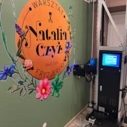 Drukarka ścienna w trakcie pracy, nanosząca kolorowy wzór z kwiatami i logo 'Natalia Czyż Warsztat Fryzjerski' na zieloną ścianę, widoczny panel sterowania drukarki.