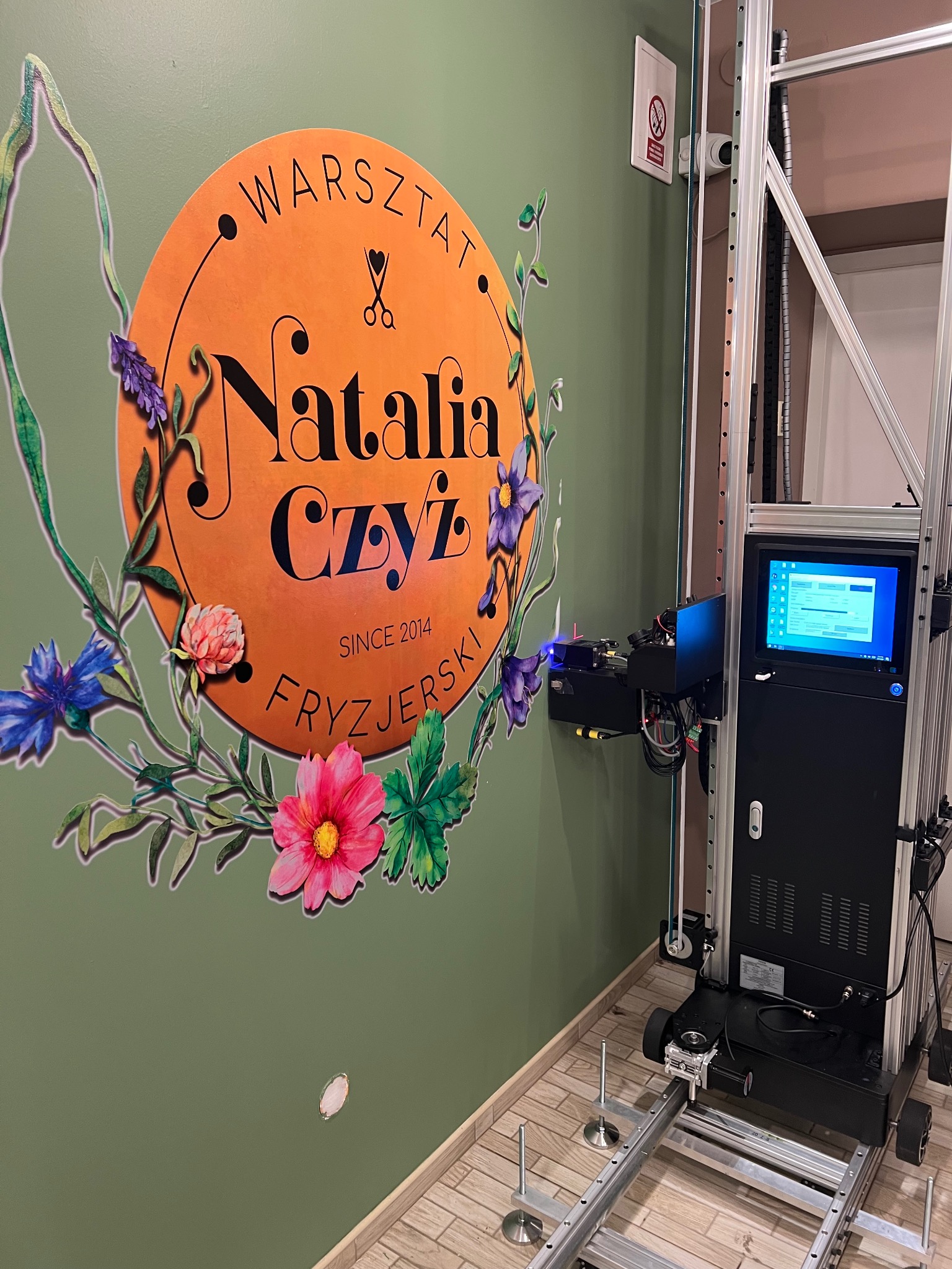 Drukarka ścienna w trakcie pracy, nanosząca kolorowy wzór z kwiatami i logo 'Natalia Czyż Warsztat Fryzjerski' na zieloną ścianę, widoczny panel sterowania drukarki.
