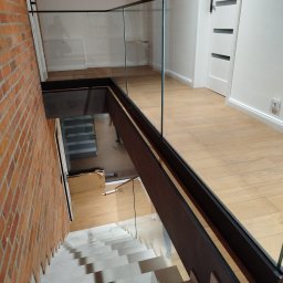 GLASS PROJECT SPÓŁKA Z OGRANICZONĄ ODPOWIEDZIALNOŚCIĄ - Szklana balustrada z czarnymi elementami mocującymi, widok z góry na schody z ceglaną ścianą. Minimalistyczny design, jasne wnętrze z drewnianą podłogą.