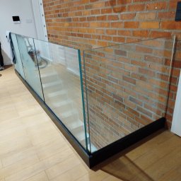 GLASS PROJECT SPÓŁKA Z OGRANICZONĄ ODPOWIEDZIALNOŚCIĄ - Szklana balustrada z czarną ramą w nowoczesnym wnętrzu z ceglaną ścianą i drewnianą podłogą. Widok z góry na schody. Minimalistyczny design.