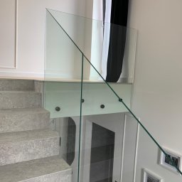 GLASS PROJECT SPÓŁKA Z OGRANICZONĄ ODPOWIEDZIALNOŚCIĄ - Szklana balustrada z mocowaniami punktowymi przy schodach z szarego kamienia. W tle okno z czarno-białą zasłoną i dekoracyjne ramki na ścianie.
