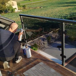 GLASS PROJECT SPÓŁKA Z OGRANICZONĄ ODPOWIEDZIALNOŚCIĄ - Osoba czyści szklaną balustradę na tarasie z widokiem na ogród. Widoczne środki czystości i narzędzia. Ujęcie z góry, w słoneczny dzień.