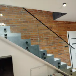 Szklana balustrada schodowa z czarnymi uchwytami na tle ceglanej ściany i białych drzwi. Minimalistyczny design, nowoczesne wykończenie.