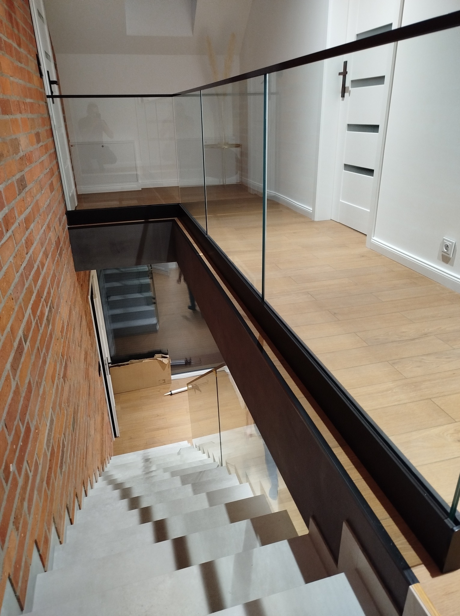 Szklana balustrada z czarnymi elementami mocującymi, widok z góry na schody z ceglaną ścianą. Minimalistyczny design, jasne wnętrze z drewnianą podłogą.
